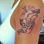Фото тату анатомического сердца, обвитого предупреждающей лентой 'CAUTION', на плече. Черно-белый рисунок татуировки выполнен в реалистичном стиле.