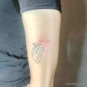 Фото тату маленького сердца с надписью "NOT YOURS" на икре ноги. Тонкий контур татуировки, выполненный черными и красными чернилами.
