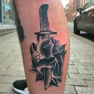 Фото тату: черно-белая татуировка в стиле blackwork на ноге. Изображено сердце, пронзенное кинжалом, с использованием плотных черных заливок.