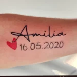 Фото тату: надпись "Amilia" с датой рождения "16.05.2020" и маленьким сердечком на руке. Минималистичный рисунок татуировки.