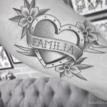 Фото тату на плече: сердце, пронзенное лентой с надписью 'Familia', в окружении цветов, выполненное в черном цвете в стиле олдскул.