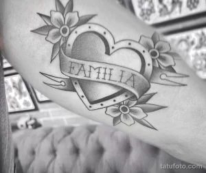 Фото тату на плече: сердце, пронзенное лентой с надписью 'Familia', в окружении цветов, выполненное в черном цвете в стиле олдскул.