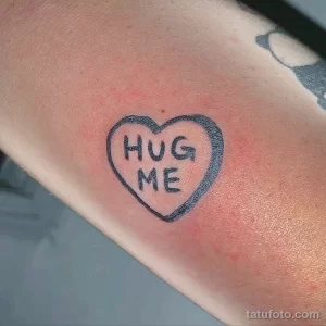 Фото тату: небольшая черно-белая татуировка в форме сердца с надписью 'Hug Me'. Простой и трогательный рисунок.