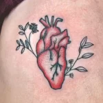 Фото тату: небольшая цветная татуировка на коже, изображающая анатомическое сердце, окруженное цветами и листьями. Милый и романтичный рисунок.