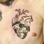 Фото тату: детализированная татуировка сердца, внутри которого изображен пейзаж с людьми, выполненная на груди. Реалистичный и сложный рисунок.