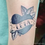 Фото тату: реалистичный рисунок граната с зелеными листьями и лентой с надписью 'LEIA'. Татуировка выполнена в стиле олдскул и расположена на предплечье.