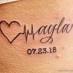 Фото тату на коже: надпись "Mayla" с датой "07.23.18" и изображением сердца, стилизованного под кардиограмму. Минималистичный и трогательный рисунок татуировки.
