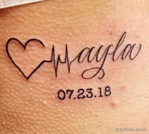 Фото тату на коже: надпись "Mayla" с датой "07.23.18" и изображением сердца, стилизованного под кардиограмму. Минималистичный и трогательный рисунок татуировки.