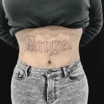 Фото тату: надпись "Angel" на талии женщины, выполненная элегантным готическим шрифтом. Черная татуировка с четкими линиями.