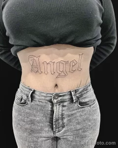 Фото тату: надпись "Angel" на талии женщины, выполненная элегантным готическим шрифтом. Черная татуировка с четкими линиями.