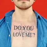 Фото тату на груди: надпись "DO YOU LOVE ME?" выполненная в стилизованном шрифте с трещинами и эффектом старения. Провокационный и эмоциональный рисунок татуировки.