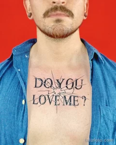 Фото тату на груди: надпись "DO YOU LOVE ME?" выполненная в стилизованном шрифте с трещинами и эффектом старения. Провокационный и эмоциональный рисунок татуировки.
