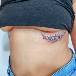 Фото тату на боку: изящная композиция из черных цветов и листьев, выполненная в графическом стиле. Элегантный и женственный рисунок татуировки.
