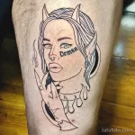 Фото тату на ноге: Изображение женского лица с рогами и надписью "Demon". Черно-белый рисунок татуировки в стиле графика.