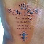 Фото тату на боку: Надпись "Mãe Pai", корона, ангельское крыло и крест. Черно-белый рисунок татуировки с религиозным подтекстом.