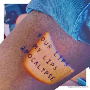 Фото тату: надпись "YOUR LIPS MY LIPS APOCALYPSE" черным цветом на руке. Небрежный стиль написания, создающий эффект рукописного текста.