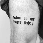 Черно-белое фото татуировки на бедре с надписью 'satan is my sugar daddy' выполненной готическим шрифтом. Ироничный рисунок татуировки для смелых личностей.