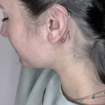 Фото тату: маленькая татуировка в виде музыкальной ноты за ухом. Черно-белый рисунок, минималистичный стиль, изящный дизайн.