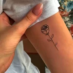 Фото тату: изящная однолинейная татуировка розы на женском запястье. Черный контур, минималистичный дизайн, нежный рисунок татуировки.
