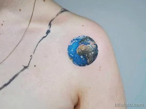 Небольшая, реалистичная татуировка на плече с изображением земного шара. Рисунок выполнен в ярких цветах и выглядит очень аккуратно.