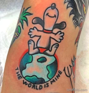 Татуировка Снупи, обнимающего Землю, с надписью "The World is Mine". Яркий рисунок в стиле old school на предплечье, символизирующий любовь к жизни.