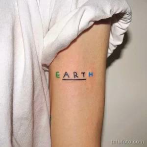Фото тату на предплечье с надписью 'EARTH', выполненной разноцветными буквами. Минималистичный рисунок татуировки с подчеркнутой линией.