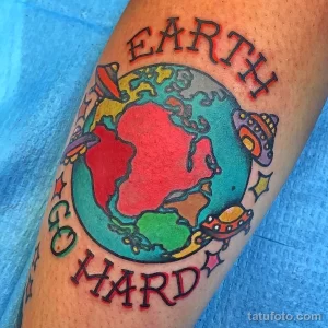 Фото татуировки на ноге с изображением Земли, НЛО, звезд и надписи "Earth Go Hard". Яркие цвета, мультяшный стиль.