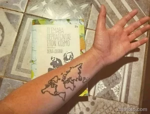 Фото тату: минималистичная карта мира на руке, рядом с книгой на греческом языке, выполненная тонкими линиями.