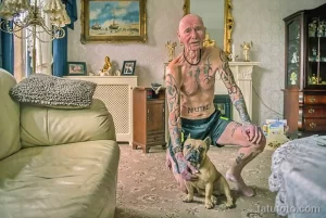 Фото тату: пожилой мужчина с татуировками обнимает французского бульдога. Яркие татуировки на теле, уютная домашняя обстановка.