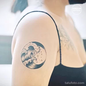 Фото тату: минималистичный рисунок волны в круге, выполненный в технике blackwork на плече девушки. Изящный эскиз тату для любителей моря.