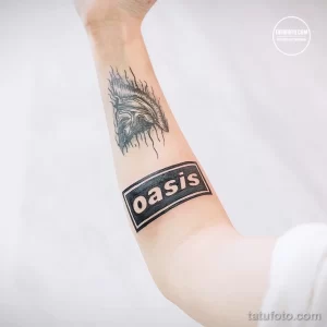 Фото тату на запястье: логотип британской группы Oasis, выполненный в черно-белом стиле. Детализированный рисунок татуировки.