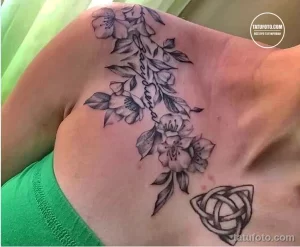 Фото тату на плече с изображением нежных цветов и надписи 'Alessia', выполненных в тонкой графике. Черно-белая татуировка.