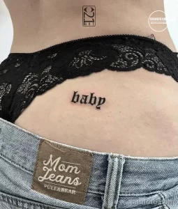Фото тату надпись 'baby' на пояснице девушки. Простой шрифт, черные чернила, минималистичный стиль. Идея для татуировки.