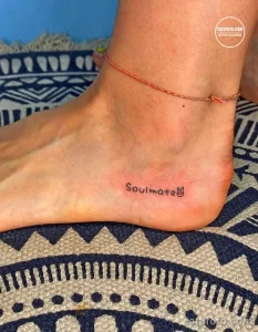 Фото тату на лодыжке: надпись "Soulmate" с изображением милого мультяшного медвежонка, выполненная тонкими линиями.