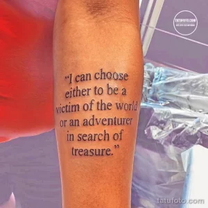 Фото тату на руке с надписью "I can choose either to be a victim of the world or an adventurer in search of treasure.". Минималистичный рисунок татуировки выполнен черным цветом.