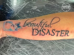 Фото тату на предплечье с надписью "Beautiful Disaster" и изображением бабочки в рамке. Черно-серый рисунок, выражающий противоречивые чувства.
