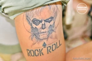 Фотография татуировки черепа с агрессивным выражением лица и надписью 'Rock and Roll' на руке. Стильный рисунок татуировки для любителей музыки.