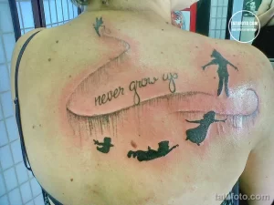 Фото тату: Цветная татуировка с надписью "Never Grow Up", изображением Питера Пэна и силуэтами летающих детей. Игривый и ностальгический дизайн.