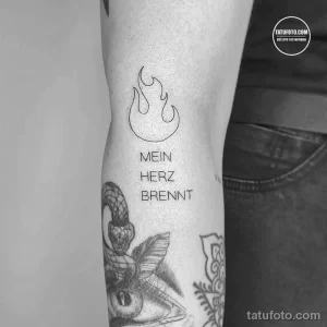 Фото татуировки в виде пламени над надписью "MEIN HERZ BRENNT" (Мое сердце горит) на руке. Минималистичный дизайн в черном цвете.