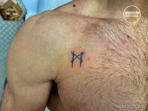 Фото минималистичной татуировки рунического символа на ключице. Тонкие линии, простой дизайн.