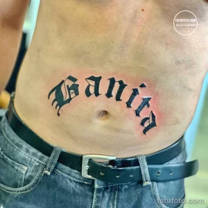 Фото тату надписи 'Vanita' выполненной в готическом стиле черными чернилами на животе. Эскиз татуировки подчеркивает индивидуальность и стиль.