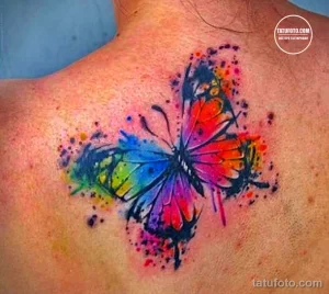 Фото тату: красочная акварельная бабочка, выполненная в технике splash ink, на женской груди. Оригинальный и яркий рисунок.