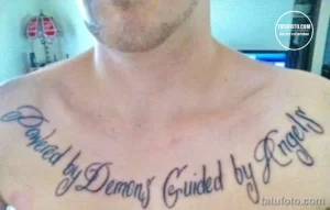 Фото тату на ключицах с надписью 'Powered by Demons, Guided by Angels'. Надпись выполнена каллиграфическим шрифтом в черном цвете.