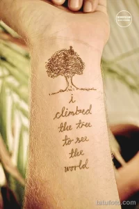 Фото тату на предплечье с изображением дерева и надписи 'i climbed the tree to see the world'. Минималистичный стиль, коричневые тона.