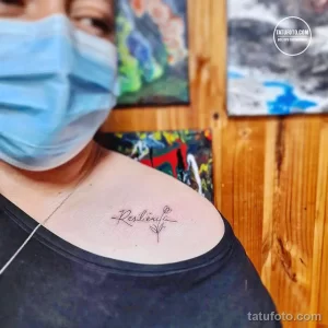 Фото тату: изящная надпись 'Resilience' с небольшими цветочными элементами, выполненная на груди женщины. Минималистичный стиль.