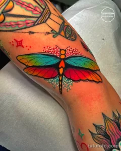Фото тату: большая стрекоза с яркими, разноцветными крыльями на руке. Татуировка выполнена в стиле акварель с использованием множества оттенков.