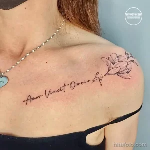 Фото тату на ключице с изображением цветка магнолии и надписью 'Amor Vincit Omnia' (Любовь побеждает все). Нежный и изящный рисунок.