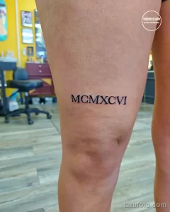 Черно-белое фото тату с римскими цифрами MCMXCVI на внутренней стороне бедра. Минималистичный дизайн, четкие линии, современный стиль.