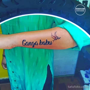 Фото тату: минималистичная татуировка с надписью "Ganga babu" и изображением летящей птицы на руке мужчины.