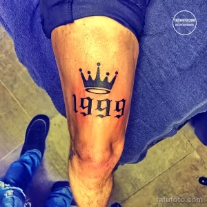Черно-белое фото татуировки на ноге, изображающей корону и год рождения '1999'. Простой и лаконичный дизайн.